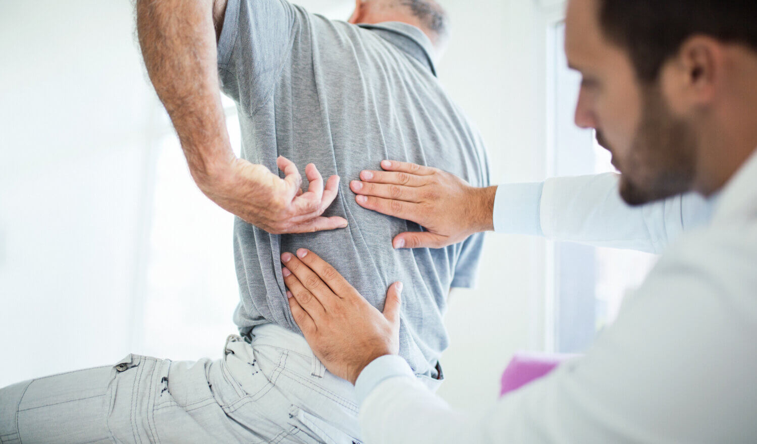 Closeup rear low angle view of a senior gentleman having some back pain. He's at doctor's office having medical examination by a male doctor. The patient is pointing to his lumbar region.