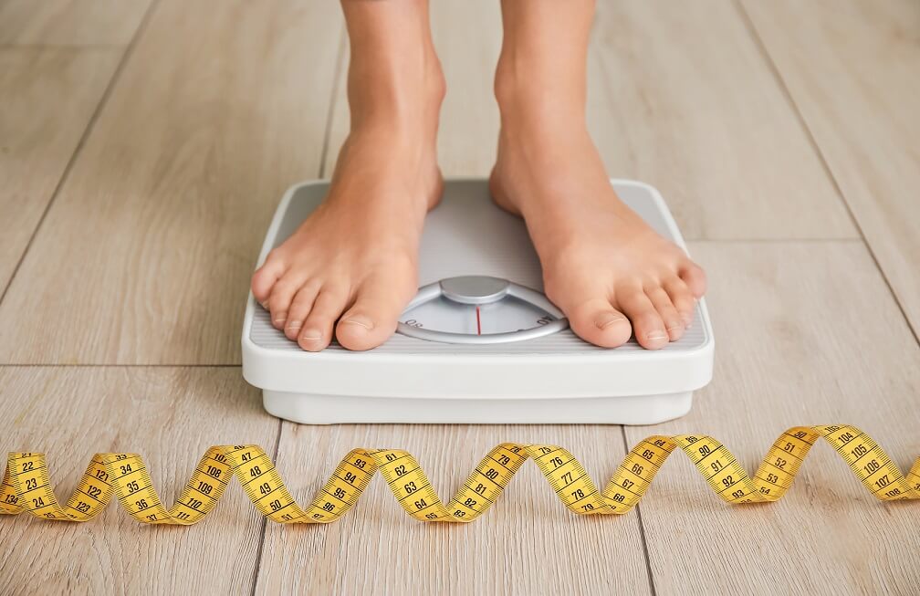 Feet standing on bathroom scale with tape measure coiled on the ground in foreground