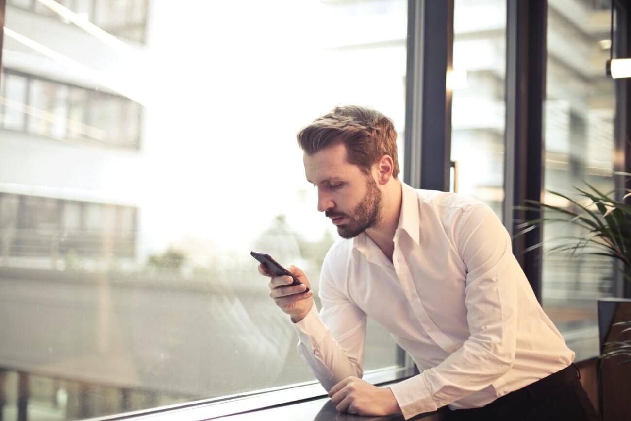 Young man next to window in office building looking down at mobile phone