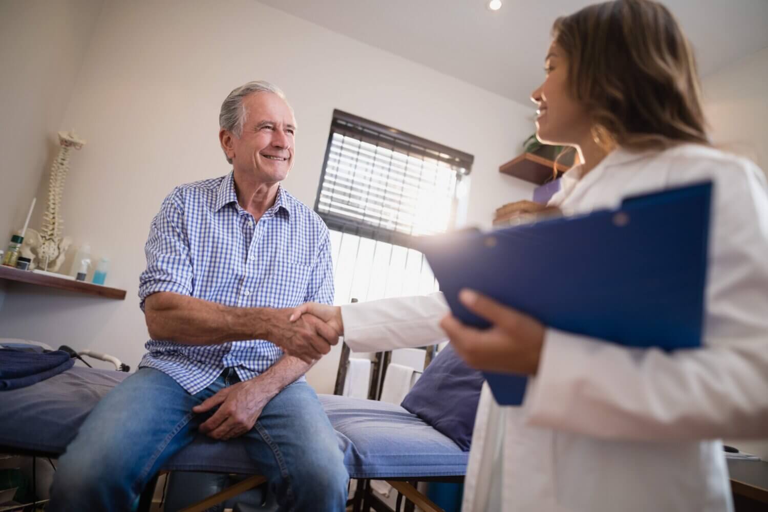 Young female doctor shaking hands with a senior male patient who is sitting on an exam table